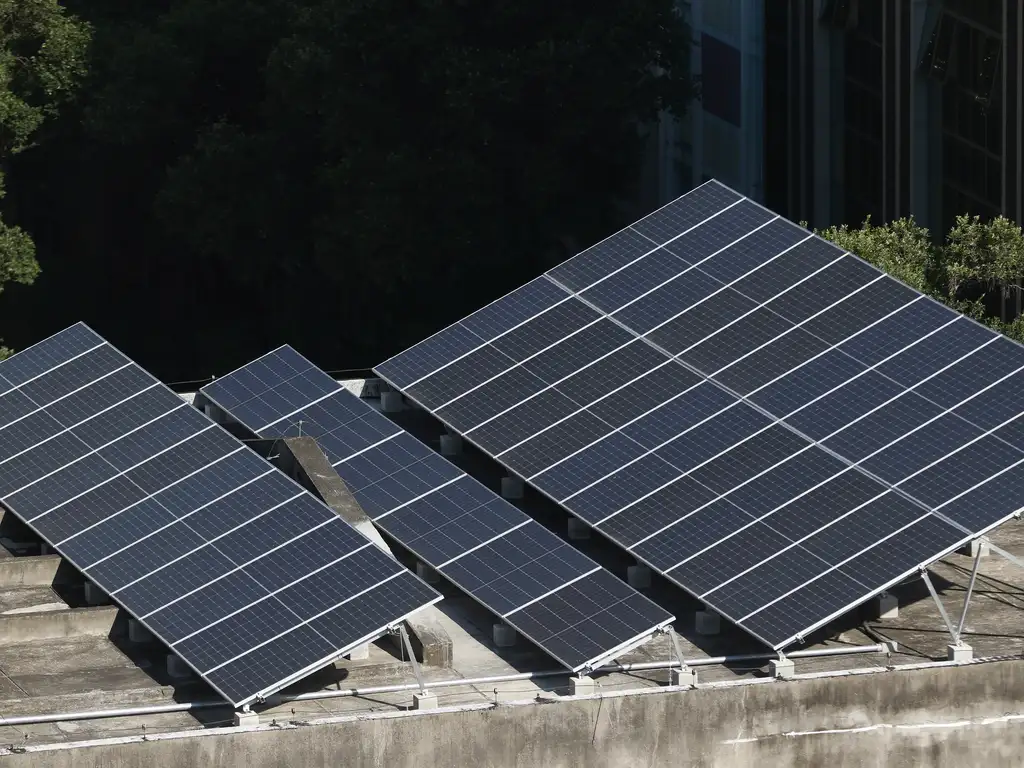 A imagem mostra um conjunto de painéis solares instalados sobre um telhado de concreto. Os painéis estão inclinados, alinhados em fileiras e voltados para a mesma direção, captando a luz solar. Ao fundo, há árvores e parte de um prédio, sugerindo que o sistema está em uma área urbana com vegetação próxima. A cena foi fotografada em um dia claro, com boa iluminação natural.