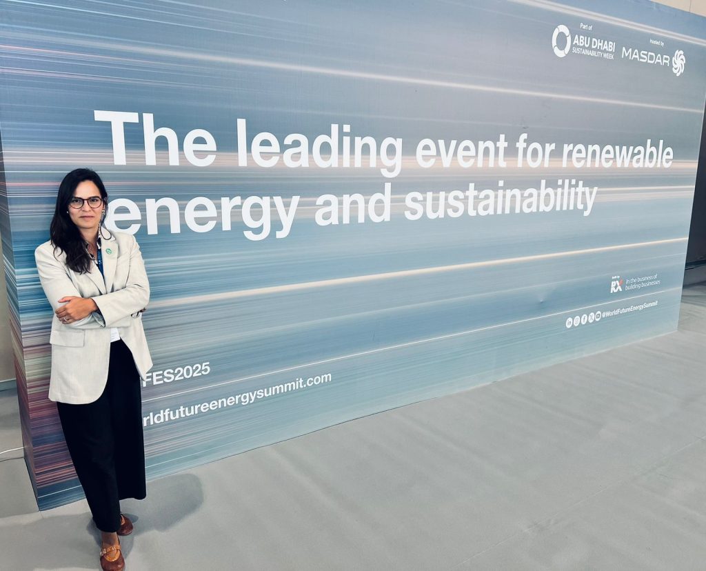 A imagem mostra Thaynara Leal em pé, de braços cruzados, posando diante de um grande painel de fundo azul e cinza com faixas de luz horizontais. O painel exibe o texto em inglês “The leading event for renewable energy and sustainability” (“O principal evento sobre energia renovável e sustentabilidade”) e menciona que faz parte da Abu Dhabi Sustainability Week, organizado pela Masdar. Thaynara veste blazer claro, calça preta e sapatos marrons, e usa óculos. O ambiente é iluminado e tem aparência moderna, típico de um centro de convenções.