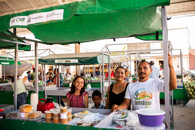 A imagem mostra uma cena em uma feira livre ao ar livre. Uma família de quatro pessoas está reunida atrás de uma barraca de mercado coberta por um toldo verde e branco com as palavras CAR e Bahia. A barraca exibe diversos produtos alimentícios, como potes de conservas, itens embalados e produtos de panificação. Um dos adultos veste uma camiseta branca com o logotipo Semiárido Show, sugerindo que o evento pode estar relacionado a uma iniciativa regional voltada para agricultura ou desenvolvimento comunitário. Ao fundo, é possível ver outras barracas e vendedores, compondo um ambiente movimentado e acolhedor, típico de feiras locais que valorizam o comércio de produtos regionais e a participação familiar.