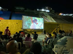 Um grupo de moradores, incluindo crianças e adultos, assiste a uma sessão de cinema ao ar livre à noite. Eles estão sentados em cadeiras plásticas, voltados para uma grande tela e para uma projeção no muro de arrimo que exibe um filme.