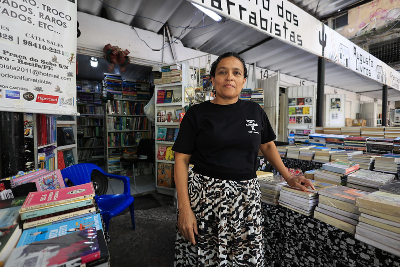 A foto mostra Kátia Sales, uma mulher de pele clara, cabelos escuros presos e expressão serena, em pé dentro de uma banca de livros localizada em um mercado popular. Ela veste uma camiseta preta com letras brancas e uma saia estampada. Ao seu redor, há pilhas e prateleiras repletas de livros usados e raros, criando um ambiente acolhedor e culturalmente rico. Acima de sua cabeça, um cartaz informa os serviços oferecidos — compra, venda e troca de livros — além de seus contatos e redes sociais. O espaço tem estrutura simples, com teto de metal ondulado e uma cadeira plástica azul ao fundo.