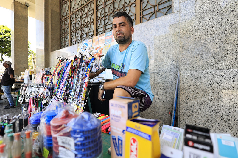 A foto mostra Cleyton Brilhante, um homem de pele morena, cabelo curto e expressão tranquila, sentado em um banquinho atrás de uma mesa repleta de materiais escolares e de escritório. Ele veste uma camiseta azul clara e shorts listrados. A mesa está organizada com itens como canetas, lápis, borrachas, corretivos, cordões de crachá e cadernos, todos dispostos de forma acessível para os clientes. Ao fundo, há uma parede de pedra com uma grade metálica decorativa, e o ambiente é parte de uma movimentada feira de rua, com outras barracas e pessoas circulando.