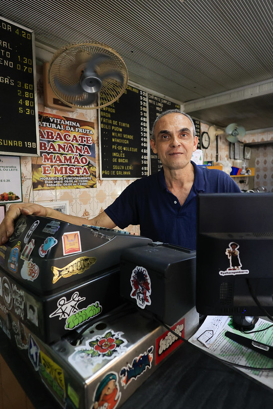 Foto de José Pinheiro sorridente atrás do balcão de sua lanchonete, vestindo uma camisa azul escura. O balcão está decorado com adesivos coloridos de personagens e logotipos, transmitindo um clima descontraído e acolhedor. Ao fundo, há dois quadros de menu com opções de sucos naturais — como uva, laranja e maracujá — e pratos variados à base de bife, incluindo versões aceboladas, à milanesa e à parmegiana. Um cartaz também destaca vitaminas feitas com frutas como abacate, banana e mamão. A parede tem um padrão geométrico discreto e um ventilador está ligado acima dos menus.