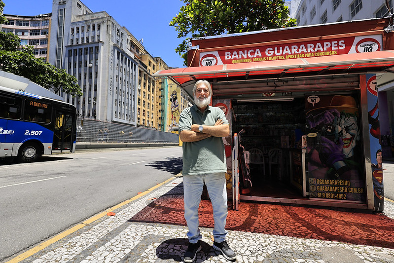 Foto colorida mostra Orlando Patrocínio em pé, de braços cruzados, em frente à tradicional Banca Guararapes, localizada na Avenida Guararapes, no centro do Recife. Ele é um homem de barba e cabelo grisalhos, veste camiseta verde e calça clara. A banca tem fachada vermelha com letreiro amarelo que anuncia a venda de quadrinhos, jornais, revistas e apostilas para concursos. Nas laterais, há ilustrações de personagens de histórias em quadrinhos, como o Coringa e o Superman. Ao fundo, aparecem prédios antigos da avenida e um ônibus passando pela rua. A cena foi registrada em um dia ensolarado.
