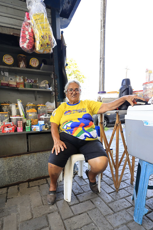 A foto mostra Maurinete Inácio, uma pessoa idosa de pele clara, sentada confortavelmente em um banquinho branco diante de sua pequena banca de rua. Ela veste uma camiseta amarela estampada em azul, shorts pretos e sapatos marrons. Com uma expressão tranquila, Maurinete apoia um braço em um isopor e o outro no banco. Atrás dela, há um quiosque simples repleto de produtos variados: pacotes de biscoitos pendurados, potes e caixas com alimentos e bebidas organizados em prateleiras. O chão é de tijolos e há um suporte de madeira ao lado do isopor.