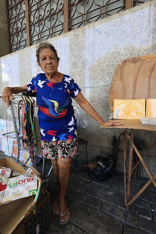 A foto mostra Terezinha Maria Camelo, uma senhora de pele clara e cabelos grisalhos, em pé ao lado de sua banca improvisada em uma calçada urbana. Ela veste uma blusa azul vibrante com estampa de pássaros e uma bermuda florida, transmitindo alegria e personalidade. Sua banca é composta por um carrinho metálico com cordões coloridos pendurados, uma caixa de papelão com bebidas embaladas — possivelmente suco de maçã — e uma mesa de madeira com pacotes amarelos organizados em pilhas. Ao fundo, vê-se uma parede de pedra com janelas gradeadas.