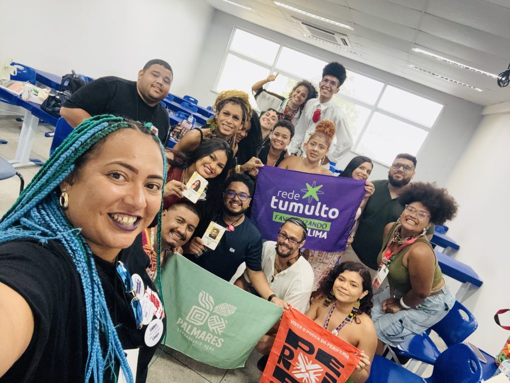 A imagem mostra um grupo de pessoas reunidos, segurando faixas, com frases: Rede Tumulto, favelizando o clima Palmares, laboratório-ação e viver o poder da periferia.