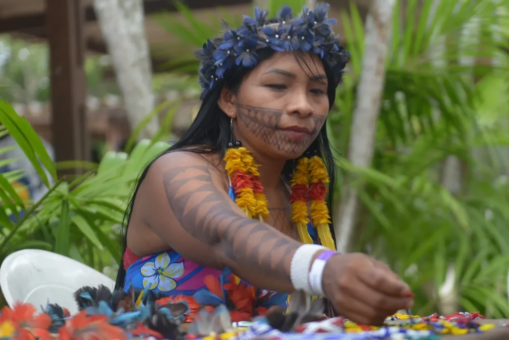 Na imagem, a indígena Gilmara Munduruku aparece sentada, envolvida em uma atividade artesanal, cercada por penas coloridas e miçangas sobre uma mesa. Ela veste um vestido florido e adornos feitos com flores vermelhas e amarelas, além de uma tiara de flores azuis. Seu rosto e braço estão pintados com grafismos pretos em padrões geométricos, típicos da cultura Munduruku. O cenário é ao ar livre, com vegetação verde ao fundo e uma estrutura de madeira, sugerindo um ambiente comunitário em meio à natureza.