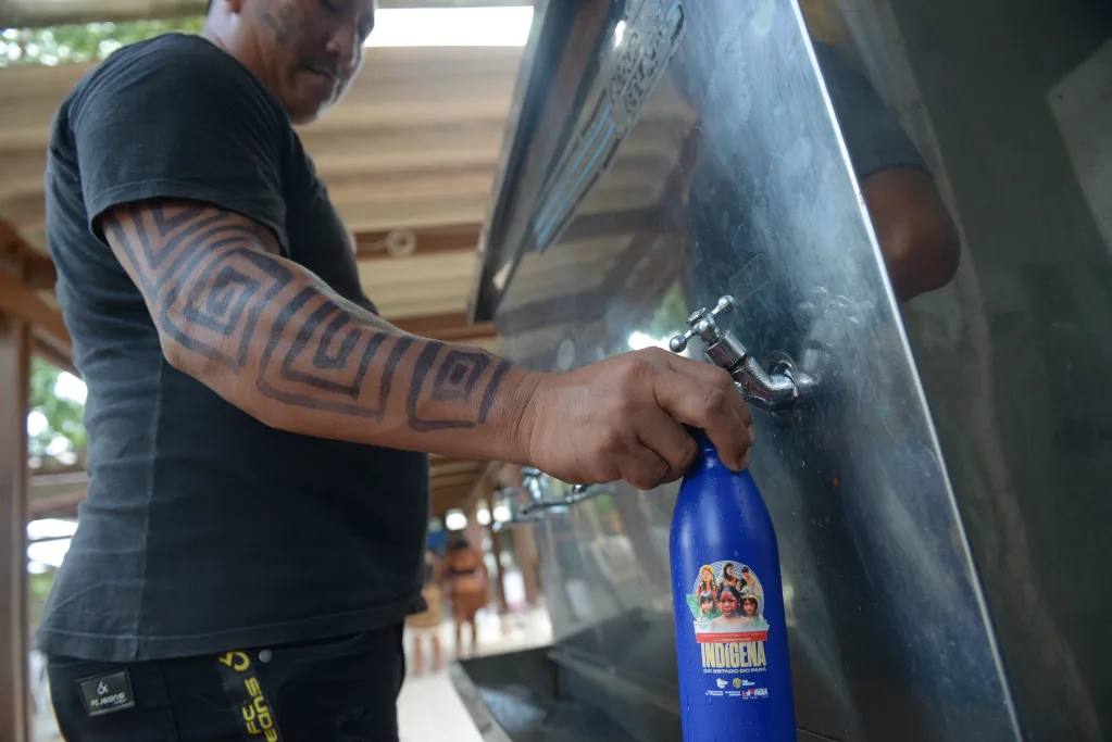 Na imagem, uma pessoa está enchendo uma garrafa azul reutilizável com a palavra “INDÍGENA” escrita em destaque, usando um bebedouro público metálico com várias torneiras. Ela veste camiseta preta e shorts pretos com letras amarelas, e tem uma tatuagem geométrica marcante no braço. Ao fundo, vê-se uma estrutura de madeira com telhado e outras pessoas circulando, sugerindo um ambiente comunitário ao ar livre.