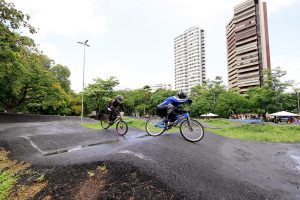 Três ciclistas equipados com capacetes e roupas de proteção percorrem uma pista de BMX com curvas e pequenas elevações, demonstrando agilidade e controle sobre suas bicicletas. O cenário é um parque urbano cercado por áreas verdes e prédios altos ao fundo, sugerindo uma localização em meio à cidade. O chão está úmido, indicando que choveu recentemente, e o céu parcialmente nublado reforça essa impressão. Ao fundo, há tendas montadas e pessoas reunidas, possivelmente espectadores ou participantes de um evento esportivo.