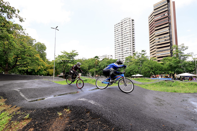 Três ciclistas equipados com capacetes e roupas de proteção percorrem uma pista de BMX com curvas e pequenas elevações, demonstrando agilidade e controle sobre suas bicicletas. O cenário é um parque urbano cercado por áreas verdes e prédios altos ao fundo, sugerindo uma localização em meio à cidade. O chão está úmido, indicando que choveu recentemente, e o céu parcialmente nublado reforça essa impressão. Ao fundo, há tendas montadas e pessoas reunidas, possivelmente espectadores ou participantes de um evento esportivo.