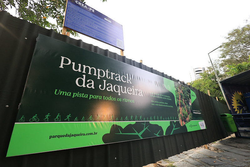 A imagem mostra uma placa verde instalada em uma cerca preta, promovendo o Pumptrack da Jaqueira, com o slogan Uma pista para todos os ritmos. O espaço é voltado para atividades como bicicleta, skate e patinete, indicado por ícones ilustrativos. À direita da placa, há uma imagem aérea da pista com curvas e elevações suaves. O ambiente ao redor é arborizado, com prédios ao fundo, sugerindo um parque urbano. Na parte inferior da placa, aparece o site parquedajaqueira.com.br, indicando que o local faz parte do Parque da Jaqueira.
