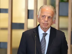 Foto de José Múcio Monteiro em um ambiente fechado, à frente de uma parede de cor marrom clara, provavelmente de lambris de madeira. Ele é um homem branco, idoso, usando paletó preto, de camisa branca e gravata cinza escura.