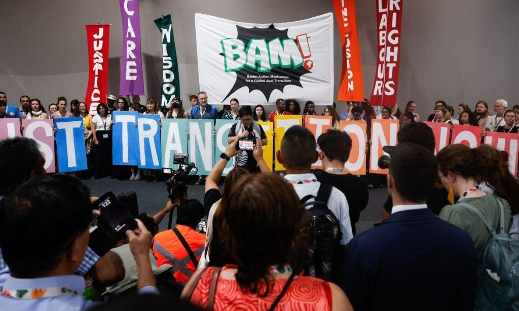 A imagem mostra um evento coletivo realizado em ambiente interno, com muitas pessoas reunidas em apoio à justiça climática e econômica. No centro, uma pessoa fala ao microfone, enquanto outras seguram grandes letras coloridas que formam a frase “JUST TRANSITION NOW”. Atrás delas, há banners verticais com palavras como “JUSTICE”, “CARE”, “WISDOM”, “VISION”, “LABOUR RIGHTS” e “SOLIDARITY”, destacando os valores defendidos pelo grupo. Um dos cartazes traz a sigla “BAM!” acompanhada da frase “Belém Action Movement for Climate and Economic Justice”, indicando a presença de movimentos racializados. O público é diverso, com pessoas de diferentes idades e origens, demonstrando engajamento e atenção ao discurso.