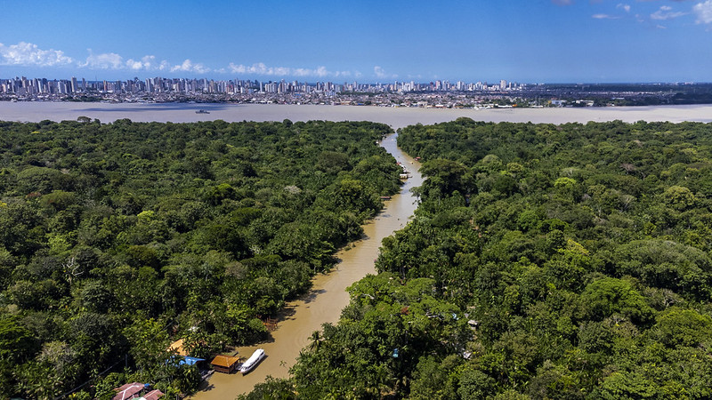 A imagem mostra uma floresta tropical densa cortada por um rio sinuoso de águas barrentas, possivelmente devido a sedimentos ou chuvas recentes. Às margens do rio, há pequenas embarcações e construções simples, indicando presença humana em meio à natureza. A vegetação é exuberante e cobre grande parte da paisagem, transmitindo a sensação de um ambiente vivo e preservado. Ao fundo, além de um largo corpo d’água, surge a cidade de Belém, com seu horizonte urbano marcado por prédios altos. O contraste entre o verde intenso da floresta e a verticalidade da cidade evidencia o encontro entre o natural e o urbano.