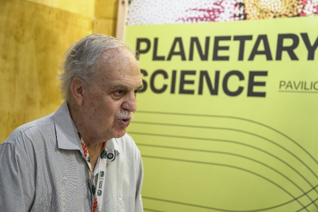 Na imagem, vemos Carlos Nobre, climatologista brasileiro, em pé diante de um painel verde com os dizeres PLANETARY SCIENCE PAVILION (Pavilhão de Ciência Planetária). Ele está vestido com uma camisa clara de manga comprida e usa um crachá pendurado no pescoço, preso por um cordão com logotipos e textos diversos, indicando sua participação oficial na COP30. O fundo inclui uma parede amarelada e parte de uma faixa decorativa com padrões.
