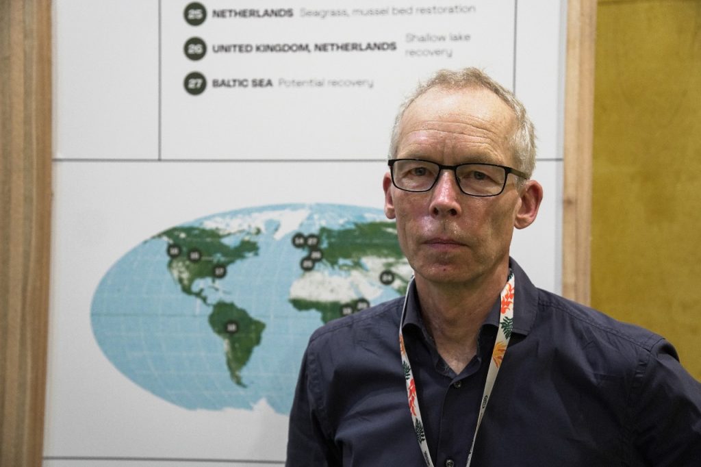 Na imagem, o cientista sueco Johan Rockström aparece em pé diante de um painel informativo que exibe um mapa-múndi com marcadores numerados e uma lista de projetos de recuperação ambiental em diferentes regiões, como a restauração de pradarias marinhas na Holanda e a recuperação do Mar Báltico. Vestido de forma discreta, ele parece participar de um evento científico voltado à sustentabilidade. Rockström é diretor do Instituto Potsdam e criador do conceito dos limites planetários, que define os limites seguros para a atividade humana no planeta.