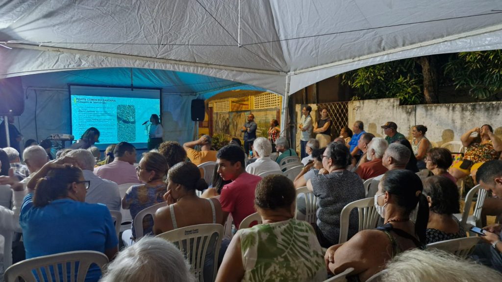 A imagem mostra uma reunião comunitária ao ar livre, sob uma grande tenda branca, com dezenas de pessoas sentadas em cadeiras plásticas, voltadas para uma tela de projeção. Algumas usam máscaras, indicando cuidados com a saúde. A apresentação exibida na tela tem o título “Projeto Urbanístico” e inclui um texto explicativo e uma imagem aérea de um bairro. O ambiente é simples, cercado por muros e vegetação, sugerindo que o evento ocorre em uma área residencial. Algumas pessoas estão em pé próximas à tela, provavelmente organizadores ou apresentadores.