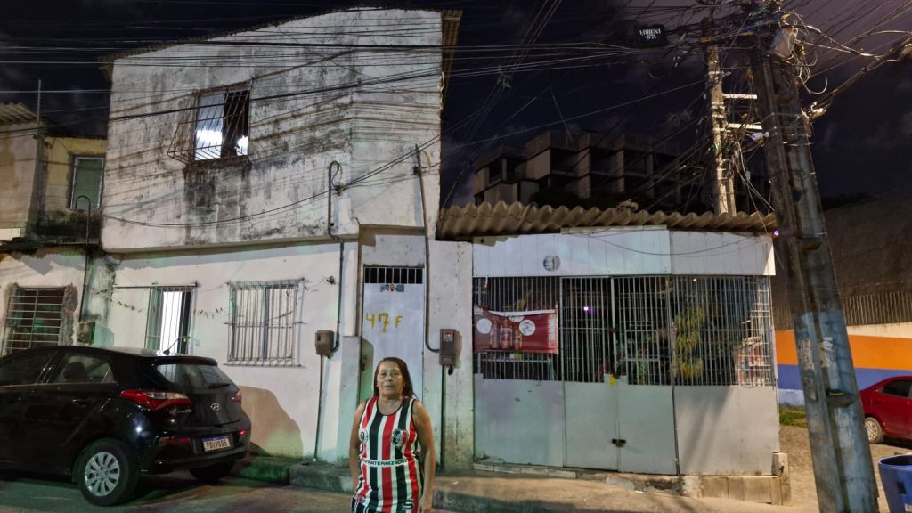 A foto mostra uma mulher em pé na calçada, à noite, em frente a uma casa simples de dois andares com paredes brancas e aparência desgastada. Ela veste uma camisa sem mangas listrada em vermelho, preto e branco, com o escudo do Santa Cruz Futebol Clube. Ao lado dela há um carro preto estacionado e, à direita, um poste com muitos fios elétricos entrelaçados. A fachada da casa tem janelas e portões com grades de ferro; na parede há o número “47F” pintado em amarelo. O ambiente é urbano e residencial, iluminado por luzes artificiais.