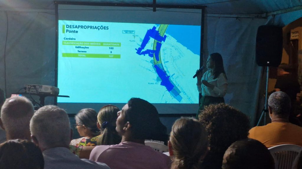 A foto mostra uma reunião pública noturna, realizada sob uma tenda. Várias pessoas estão sentadas, de costas para a câmera, assistindo a uma apresentação projetada em uma tela. Uma mulher em pé, à direita, segura um microfone e aponta para o slide, que exibe um mapa e o título “DESAPROPRIAÇÕES – Ponte”. No slide, aparecem dados sobre o bairro Cordeiro, indicando a quantidade de imóveis afetados: 102 edificações e 5 terrenos, totalizando 107. O ambiente é iluminado principalmente pela luz da projeção, e há um projetor sobre uma mesa à esquerda. A cena transmite um clima de reunião comunitária ou audiência pública para discutir intervenções urbanas.