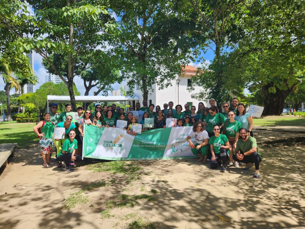 A imagem mostra um grupo de pessoas reunidas ao ar livre, em frente a um prédio branco, segurando uma faixa verde e branca com os dizeres “Maio Celíaco”. Muitos estão vestidos com camisetas verdes e alguns exibem certificados. O evento parece ser uma ação de conscientização sobre a doença celíaca. O clima é ensolarado e o ambiente transmite união e engajamento comunitário.