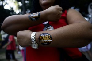 Uma pessoa exibe dois adesivos nos antebraços com frases: “Mulheres Negras movem estruturas!” e “Povo Negro Forte”. Ela veste uma blusa vermelha sem mangas e usa um relógio no pulso esquerdo. Ao fundo, há outras pessoas e árvores, sugerindo um evento ao ar livre.