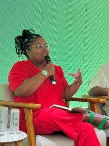 A imagem mostra uma jovem negra, com cabelos trançados presos, óculos de grau e um macacão vermelho, sentada falando ao microfone.