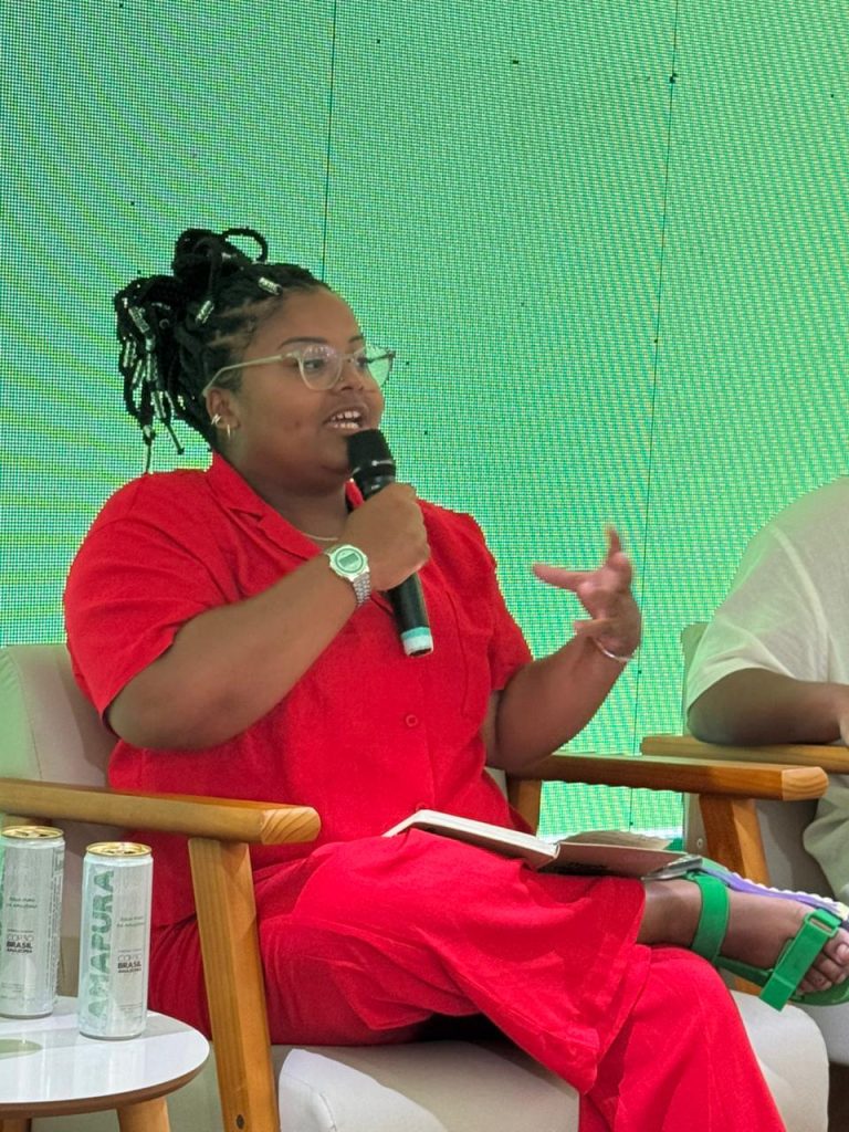 A imagem mostra uma jovem negra, com cabelos trançados presos, óculos de grau e um macacão vermelho, sentada falando ao microfone.