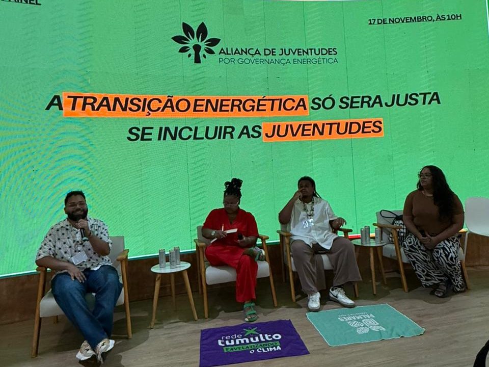 A imagem mostra um painel com quatro pessoas discutindo sobre juventude e governança energética. Ao fundo, uma tela verde exibe o tema do evento: “A transição energética só será justa se incluir as juventudes”. O ambiente é formal, com cadeiras para o público e materiais de divulgação no chão.
