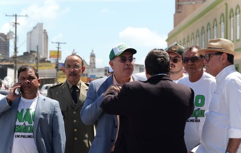 A imagem mostra um grupo de seis homens reunidos em uma rua sob céu azul e ensolarado. Eles conversam entre si, parecendo participar de um evento público ou manifestação. Dois deles vestem camisetas brancas com estampas verdes; um usa boné e óculos escuros. Outro homem, de terno preto, aparece de costas, conversando com o grupo. Um dos participantes veste uniforme que lembra o de forças de segurança. Ao fundo, aparecem prédios comerciais, calçadas e postes, indicando que a cena ocorre em uma área urbana movimentada.