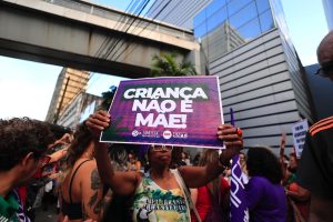 A imagem mostra uma mulher negra participando de uma manifestação segurando um cartaz com os dizeres “CRIANÇA NÃO É MÃE!”. Ao fundo, há outras pessoas e prédios urbanos. O cartaz e o contexto indicam um protesto em defesa dos direitos das crianças e das mulheres.