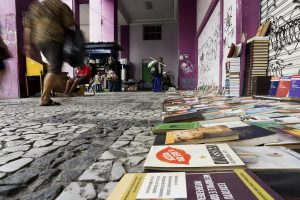 A foto mostra uma feira de livros ao ar livre, montada sobre um calçadão de pedras em um espaço urbano. À direita, há uma grande quantidade de livros dispostos diretamente no chão, com as capas voltadas para cima. Os títulos são variados, incluindo literatura, autoajuda e materiais didáticos — alguns visíveis são “Verissimo”, “Estudo Dirigido de Interpretação de Textos” e “O Papo Reto”. Próximo a uma parede roxa coberta por grafites, há uma pilha alta de livros encostada verticalmente. Ao fundo, pessoas caminham e exploram a feira, que conta com outras bancas e quiosques.