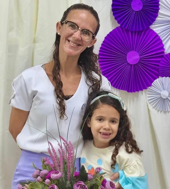 A foto mostra uma mulher adulta e uma criança posando juntas, sorrindo para a câmera, diante de um fundo decorado com círculos de papel sanfonado em tons de roxo e lilás. A mulher está em pé, usa óculos, tem cabelos longos e cacheados presos para trás, e veste uma blusa clara com mangas curtas. A criança está encostada ao lado dela, com cabelos cacheados soltos, usando uma tiara azul clara e uma blusa em tons pastel. À frente delas há um arranjo de flores roxas e verdes.