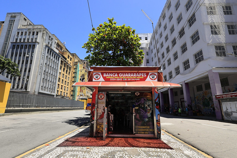 A foto mostra a Banca Guararapes em primeiro plano, no centro da Avenida Guararapes, no Recife, em um dia de céu azul. A banca tem cobertura vermelha, nome destacado na parte superior e ilustrações de quadrinhos nas laterais, com o interior parcialmente visível. Logo atrás dela, uma árvore frondosa se destaca. À esquerda, aparece um prédio cinza de arquitetura antiga, e à direita, um edifício branco parcialmente coberto por tela de obra. A calçada de pedras portuguesas e a rua vazia completam a cena urbana.