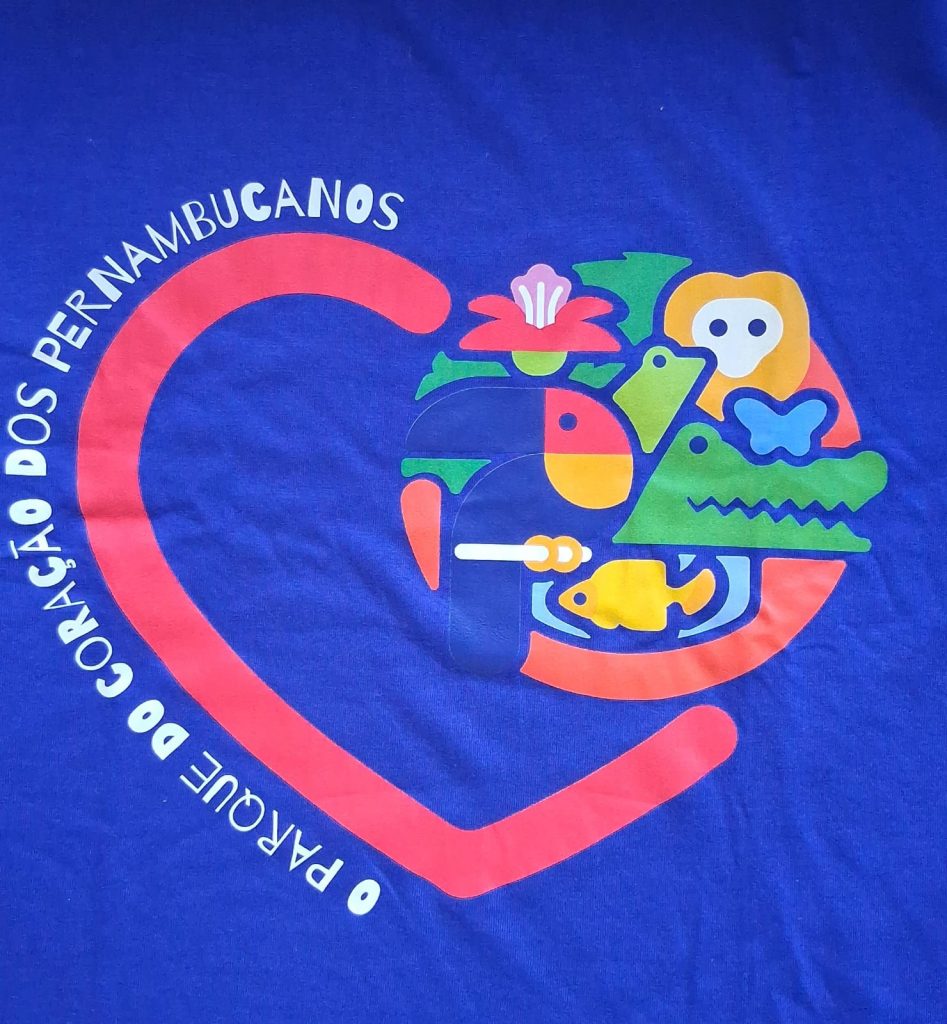 A imagem mostra uma estampa colorida sobre um fundo azul. O desenho principal é um coração formado por um traço vermelho, dentro do qual há figuras estilizadas e geométricas representando elementos da fauna e flora nordestina: um tucano, um peixe, um jacaré, uma flor, uma borboleta e um macaco. As formas são simples, coloridas em tons de verde, amarelo, azul, vermelho e laranja. Em volta do coração, está escrito em letras brancas a frase: “O PARQUE DO CORAÇÃO DOS PERNAMBUCANOS”.