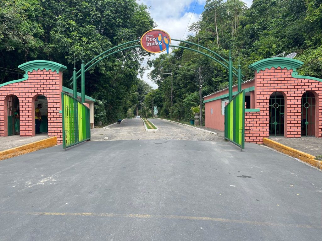 A imagem mostra a entrada principal do Parque Dois Irmãos, no Recife. O portão é formado por duas pequenas construções simétricas, feitas de alvenaria pintada em tom rosado com detalhes que imitam tijolos vermelhos e molduras verdes. Entre elas, há um grande arco de ferro verde com um letreiro circular no alto, onde se lê “Parque Dois Irmãos” sobre um fundo vermelho. Os portões metálicos, também verdes, estão abertos, revelando uma via de paralelepípedos que segue entre árvores densas de mata nativa. O céu está parcialmente nublado.