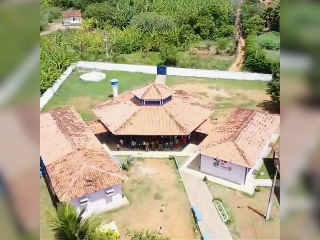 A imagem mostra, vista de cima, a estrutura da Escola Pajé Miguel Celestino, composta por vários prédios térreos com telhados de cerâmica alaranjada distribuídos ao redor de um pátio central. As construções têm formato simples e estão conectadas por caminhos cimentados. No centro há um prédio maior, com telhado em formato octogonal, onde se vê um grupo de pessoas reunido sob uma área coberta. A escola está cercada por um muro branco, e ao redor há extensa vegetação verde com árvores e plantações, além de uma estrada de terra estreita que leva ao fundo da propriedade.