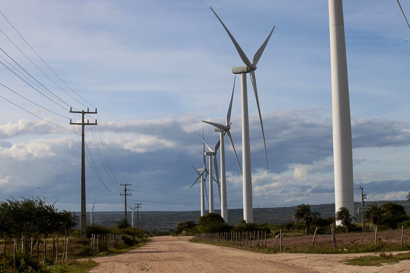 A foto mostra uma estrada de terra cercada por vegetação baixa, com vários aerogeradores (turbinas eólicas) alinhados ao lado direito e postes de energia com fios ao lado esquerdo, sob um céu parcialmente nublado. É uma paisagem rural com infraestrutura de geração e transmissão de energia.