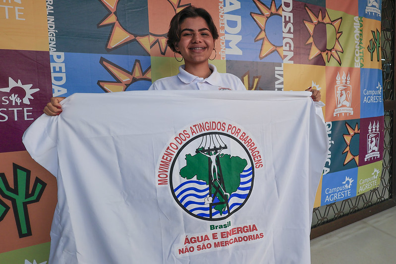 A foto mostra uma jovem branca de cabelos curtos e brincos de argola sorrindo enquanto segura uma bandeira do “Movimento dos Atingidos por Barragens”. O fundo é colorido, com vários painéis que exibem nomes de cursos e símbolos do campus da UFPE.