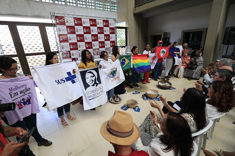 A foto mostra um grupo de pessoas alinhadas segurando diversas bandeiras e cartazes de movimentos sociais e instituições. Elas estão em um ambiente interno com várias pessoas sentadas ao redor, aplaudindo a apresentação. A cena faz parte cerimônia de abertura do curso de Medicina pelo Pronera na UFPE campus Caruaru.