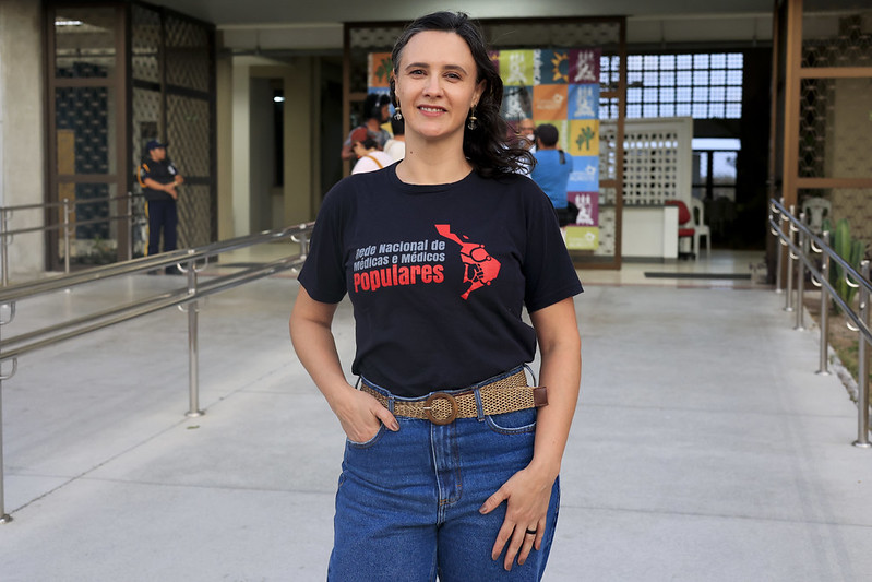 A foto mostra uma mulher branca de cabelos lisos em pé, ao ar livre, posando em frente a um prédio. Ela usa camiseta preta com estampa de um movimento de médicos populares e calça jeans, olhando para a câmera com expressão tranquila.
