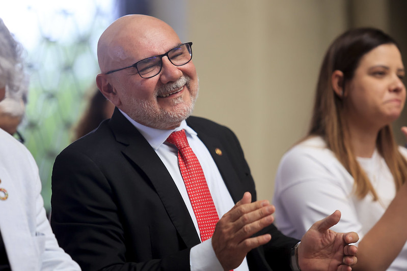 A foto mostra um homem sorrindo e aplaudindo, vestindo terno escuro, camisa branca e gravata vermelha. Ele está em um ambiente interno, acompanhado de outras pessoas ao lado.