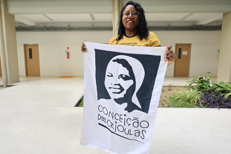 A mulher negra de cabelos cacheados e óculos de grau na foto está sorrindo e segurando uma bandeira branca com uma ilustração em preto de um rosto feminino e a frase “Conceição das Crioulas”. Ela está em um ambiente interno claro, com portas ao fundo e um pequeno jardim lateral.