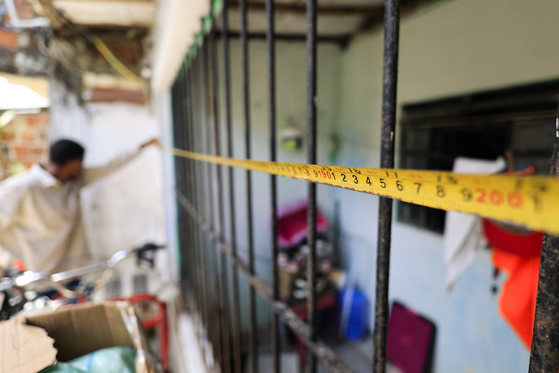 A imagem mostra uma fita métrica amarela esticada horizontalmente sobre uma grade de janela ou portão. Os números de medição estão visíveis, indo de 0 a 9 centímetros, com o número 200 também destacado, indicando uma medida maior. Ao fundo, dentro de um cômodo, há uma pessoa segurando a outra ponta da fita, e é possível ver alguns objetos domésticos como sacolas e roupas. A cena sugere que está sendo feita uma medição, possivelmente para reforma ou instalação.