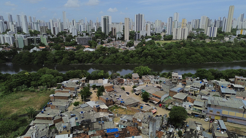 A foto mostra dois lados muito diferentes de uma cidade separados por um rio: na frente, há uma área com casas pequenas e muito próximas umas das outras, feitas com materiais simples como telhas de metal, mostrando sinais de pobreza; ao fundo, do outro lado do rio, aparecem prédios altos e modernos, indicando uma região mais rica; entre os dois lados, há uma faixa de vegetação verde que acompanha o rio, marcando a divisão entre esses espaços urbanos tão contrastantes.