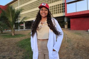 Uma jovem negra de cabelos cacheados e óculos de grau está em pé ao ar livre diante de um prédio moderno com fachada vermelha e bege. Ela veste jaleco branco, calça e blusa beges, além de um boné vermelho do MST. Há uma palmeira e vegetação seca no primeiro plano, sugerindo clima quente. O cenário parece é do campus universitário da UFPE em Caruaru.