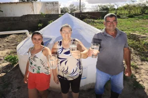 Três pessoas estão em frente a uma cisterna branca em uma área rural, sorrindo e segurando copos com água, com vegetação, casas simples e céu claro ao fundo. Há uma jovem, uma mulher e um homem adultos.