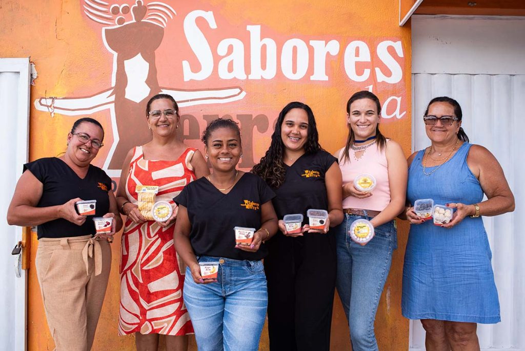 Seis mulheres que fazem parte da Associação de Mulheres abores da Terra pousando para foto. Elas seguram pequenos potes com os produtos produzidos por elas. Na parede atrás delas a logomarca da associação pintada em tons terrosos.