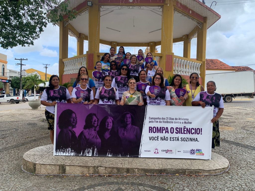 Um grupo de mulheres está reunido em frente a um coreto numa praça pública, participando de uma campanha contra a violência contra a mulher. Elas estão em pé, lado a lado, segurando cartazes e vestindo camisetas com mensagens de apoio à causa. À frente do grupo, há uma faixa grande com os dizeres: “Campanha dos 21 Dias de Ativismo pelo Fim da Violência contra a Mulher – Rompa o silêncio! Você não está sozinha.