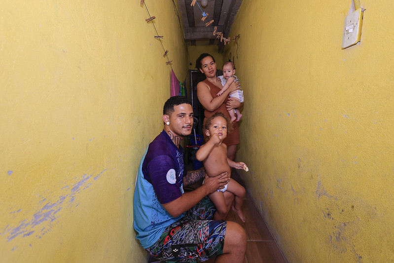 A imagem mostra um corredor estreito com paredes amarelas vivas e piso de azulejos claros, onde vive uma família composta por dois adultos e duas crianças pequenas. O teto tem vigas de concreto expostas e um fio com prendedores de roupa atravessa o espaço, sugerindo uso doméstico. Sentado no chão está Pablo Fernando, segurando um bebê com carinho, enquanto mais ao fundo uma mulher em pé segura outra criança nos braços. O ambiente é simples e apertado, mas transmite um forte senso de afeto e intimidade familiar.